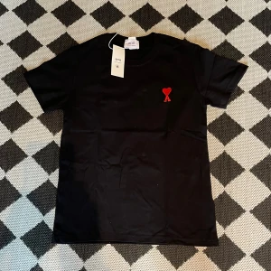 Svart Ami t-shirt  - Svart t-shirt från Ami med ett litet rött hjärta broderat på bröstet. Klassisk passform med rund hals och korta ärmar. Tillverkad i mjuk bomull för en skön känsla. Perfekt för en stilren och enkel look.