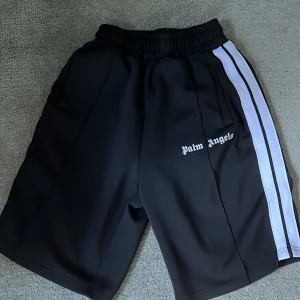 Svarta shorts med vita ränder från Palm Angels - Svarta shorts från Palm Angels med vita ränder längs sidorna och logga tryckt på vänster ben. De har elastisk midja och sidofickor. Perfekta för en sportig och avslappnad stil. Pris kan diskuteras!