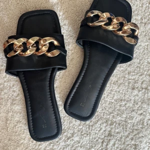Svarta sandaler med guldkedja  - Säljer ett par svarta sandaler med fyrkantig tå och bred rem framtill dekorerad med en stor guldfärgad kedja. Platt sula och öppen tå, perfekt för varma dagar. Snygg och trendig design som passar till många outfits. Köpta från dinsko 