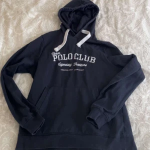 Polo club hoodie - Mörkblå hoodie med vit text på bröstet från Urban Polo Club. Tröjan har huva med vita snören och en stor magficka framtill. Perfekt för en avslappnad stil