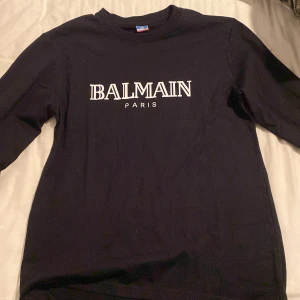 Svart långärmad tröja från Balmain Paris - Säljer en svart långärmad tröja från Balmain Paris med vit logotyp på bröstet. Tröjan har en klassisk passform och rund halsringning. Perfekt för dig som gillar stilrena och exklusiva plagg.