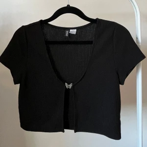 Svart croppad bolerotopp med fjärilsdetalj - Säljer en svart croppad bolerotopp från Divided. Toppen har korta ärmar, djup v-ringning och öppet snitt framtill med en söt liten fjärilsdetalj. 