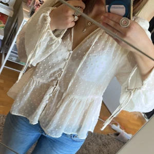 Vit blus med volanger och knytband - Säljer en söt vit blus med volanger och knytband vid halsen. Blusen har långa ärmar med resår och små prickiga detaljer i tyget. Perfekt att styla med jeans eller kjol. Storlek M, men sitter mer som en S