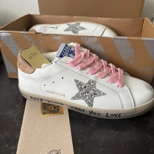 Golden Goose iconic superstar - Säljer ett par vita sneakers från Golden Goose med glittrig silverstjärna på sidan, rosa skosnören och beige hälparti. Skorna har rund tå och platt sula. Perfekta för dig som vill ha en unik och trendig look.