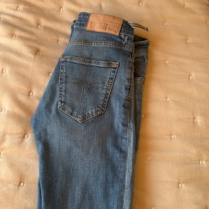 Ljusblå jeansbyxor med klassisk passform - Säljer ett par ljusblå jeans från tiger of sweden med klassiskdesign och skinnpatch i midjan. Jeansen har raka ben och normal passform, perfekta till vardags. Tidlös modell som funkar till det mesta. ett hål i dom som knappt syns när man har dom på sig därför för de priset.