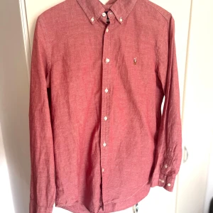 Rosa/Röd skjorta från Ralph Lauren - Säljer en klassisk röd skjorta från Ralph Lauren med button-down krage och vita knappar. Skjortan har lång ärm och en liten broderad logga på bröstet. Perfekt för dig som gillar stilrena och tidlösa plagg.