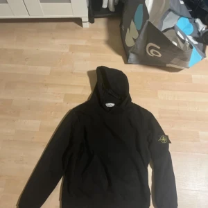 Svart hoodie från Stone Island - Säljer en svart hoodie från Stone Island med patcj på vänster ärm. Tröjan har huva och en stor magficka framtill. Perfekt för dig som gillar streetwear och vill ha något stilrent och bekvämt.