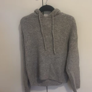 Grå stickad hoodie från Gina Tricot - Mysig grå stickad hoodie från Gina Tricot med huva och dragsko. Tröjan har lång ärm och en avslappnad passform, perfekt för kyliga dagar. Enkel och stilren design som passar till det mesta.