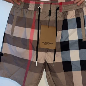Burbbery shorts - Shorts från Burberry med klassiskt rutigt mönster i beige, svart, vitt och rött. Elastisk midja med svart snörning och fickor fram och bak. Perfekta för en vanlig sommar dag men även ifall det blir bad.