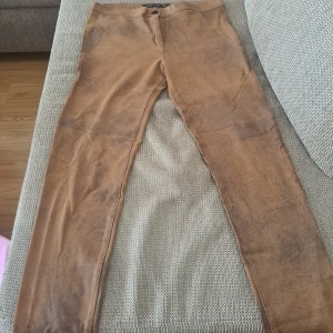 Bruna mockaimitationsbyxor - Snygga bruna byxor i mockaimitation med tight passform. Byxorna har hög midja och är utan synliga fickor eller detaljer. Perfekta för dig som gillar en stilren look med lite western-vibe.