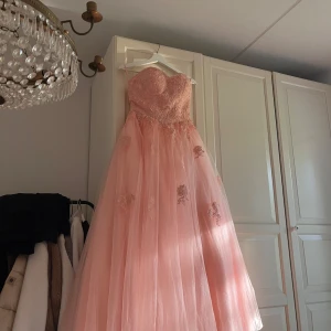 Balklänning - Jättevacker rosa/aprikos långklänning med hjärtformad överdel och vackra spetsdetaljer. Klänningen är axelbandslös och har en vid kjol i flera lager tyll. Perfekt för bal eller fest! Dragkedja bak för enkel påklädning. Aldrig använd, bara provad. Måttbeställdes för min dotters bal nu i maj men användes inte. Stl. 36 