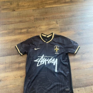 Svart Brasilien fotbollströja Stussy x Nike - Säljer en svart Brasilien fotbollströja från Nike i samarbete med Stussy. Tröjan har gulddetaljer vid krage och ärmslut, samt vit Stussy-logga på bröstet. Broderat Brasilien-emblem och Nike-märke framtill. Snygg och unik design.
