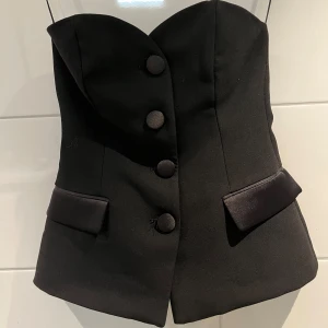 Svart korsett-topp från Zara - Säljer en svart korsett-topp från Zara med snygga stora knappar framtill och dekorativa ficklock i satin. Toppen är axelbandslös och har en figurnära passform som ger en elegant siluett. Perfekt för att lyfta vilken outfit som helst!