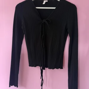 Svart ribbad knyttröja från Nelly - Säljer en svart långärmad ribbad tröja från Nelly med knyt framtill. Tröjan har croppad passform och v-ringning, perfekt till många olika outfits. Använd 2 gånger.