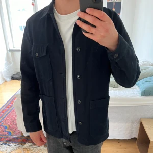 Mörkblå overshirt från A Day's March - Säljer en mörkblå overshirt från A Day's March i kraftig bomull. Jackan har knäppning framtill, tre fickor på framsidan och klassisk krage. Den saknar en knapp på ena ärmen. Extra knappar finns dock på insidan av jackan.