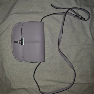 Ljuslila axelväska med silverdetalj - Säljer en stilren ljuslila axelväska med lång justerbar rem och silverfärgat spänne framtill. Väskan har flera fack inuti, inklusive ett med dragkedja, perfekt för att organisera dina saker. Enkel och snygg design som passar till många tillfällen.