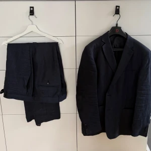 Mörkblå kostym i linne - Snygg mörkblå kostym i linne med kavaj och matchande byxor. Kavajen har klassisk passform med två knappar framtill och fickor. Perfekt för sommarens tillställningar storlek på kostymbyxor 40-32. Storlek på kavaj 58 slim fit.