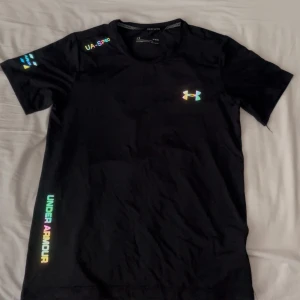Svart t-shirt från Under Armour - Säljer en svart t-shirt från Under Armour med färgglada loggor och textdetaljer på bröstet, ärmen och sidan. T-shirten är tillverkad i ett lätt och syntetiskt material som håller dig sval och torr, perfekt för träning.