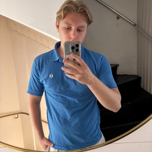 Blå piké från Fred Perry - Säljer en klassisk blå pikétröja från Fred Perry med vit broderad logga på bröstet. Tröjan har krage och korta ärmar, perfekt för en stilren och sportig look. Passar dig som gillar tidlös design och bekväma material.