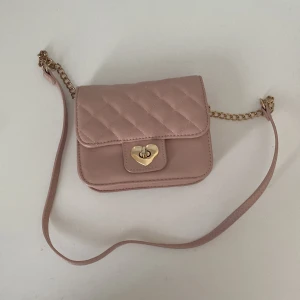 Gullig rosa axelväska med guldkedja och hjärtformad lås - Original priset runt 300-500!! Super söt ljusrosa axelväska med quiltat mönster och guldfärgad kedja! Väskan har ett hjärtformat lås framtill och ett rymligt fack inuti. Perfekt accessoar för att lyfta din outfit med en romantisk touch. Köptes i glitter.