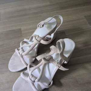 Beigea skor med klack från H&M - Säljer ett par beiga/rosa skor från H&M med hög klack och öppen tå. Skorna har snygga knutdetaljer och justerbart spänne vid ankeln. Perfekta för sommaren och ger en stilren look till din outfit. Använda en gång. 