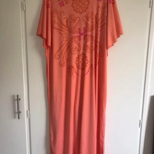 Korallfärgad koftan med mönster - Säljer en korallfärgad koftan med vida ärmar och dekorativt mönster i rosa och lila nyanser över bröstet och axlarna. Klänningen har en lös passform och är perfekt för varma dagar.