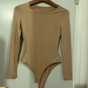 Brun långärmad bodysuit - Säljer en enkel och stilren brun bodysuit med lång ärm och rund halsringning. Perfekt att styla till jeans eller kjol för en clean look.