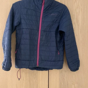 Blå pufferjacka från Norrøna - Säljer en blå pufferjacka från Norrøna med rosa dragkedja och detaljer. Jackan har lång ärm, hög krage och dragsko nertill. Perfekt för kyligare dagar och enkel att matcha med andra plagg.