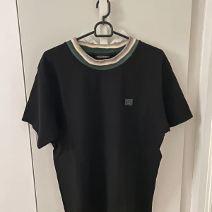 Acne studios Svart t-shirt med kontrastkrage - Säljer en svart t-shirt från Acne Studios med korta ärmar och en unik krage i grönt och beige. Klassisk passform och diskret Acne Studios-logga på bröstet. Perfekt för dig som gillar stilrena plagg med en twist. Oversized fit, köpte från acne archive i Stockholm 