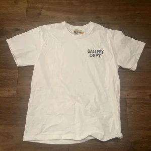 Vit t-shirt från Gallery Dept. - Vit t-shirt från Gallery Dept. med tryck på bröstet och stor text på ryggen. Klassisk passform och rund halsringning. Perfekt för en clean och stilren look. Passar mer som M