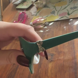 Gröna solglasögon med fjärilsform - Snygga solglasögon med grön båge och stora, lätt fjärilsformade glas. Bågarna har en silverfärgad detalj vid gångjärnet. Glasen är mörka och ger en cool retrovibe. Perfekt accessoar för att sticka ut i mängden.