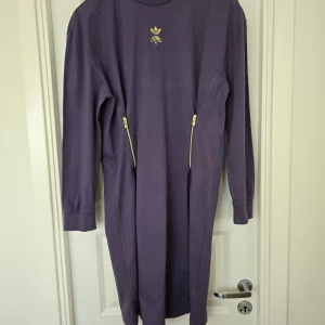 Lila långärmad klänning från Adidas - Säljer en lila långärmad klänning från Adidas med guldfärgade dragkedjor i midjan och Adidas-logga framtill. Klänningen har en rak passform och är tillverkad i mjukt bomullsmaterial. Perfekt för en sportig och avslappnad stil.