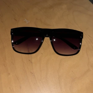 Helt nya Svarta solglasögon med fyrkantig form - Helt nya Stilrena svarta solglasögon med breda skalmar och fyrkantiga bågar. Glasen är mörka och tonade, vilket ger en cool look. Passar dig som gillar en modern och markerad stil.