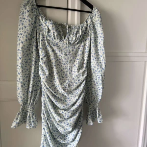 Blommig långärmad klänning från Adlore - Söt blommig klänning från Adlore med ljusblå och vita toner. Klänningen har långa ärmar med volang vid ärmslut, rynkade detaljer framtill och knytning vid halsen. Perfekt för vår och sommar.