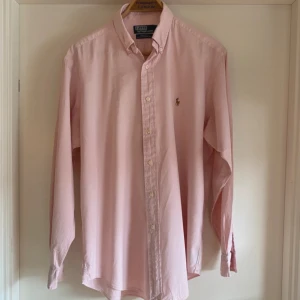 Ljusrosa skjorta från Polo Ralph Lauren - Klassisk ljusrosa skjorta från Polo Ralph Lauren med button-down krage och broderad logga på bröstet. Skjortan har långa ärmar och knäppning framtill. Perfekt för en stilren och avslappnad look.