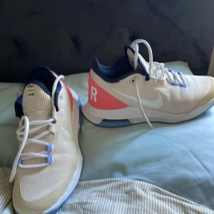 Nike springskor i vitt och korall - Säljer ett par fräscha Nike springskor i vitt med korallröda och blå detaljer. Skorna har snörning, vit sula och blå insida. De är aldrig använda och lite små i storlek, hör gärna av dig vid frågor och funderingar!