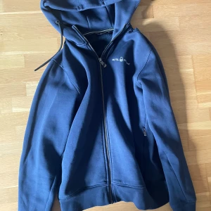 Blå hoodie med dragkedja från Sail racing - Säljer en blå hoodie från Sail racing med dragkedja framtill och huva med dragsko. Tröjan har fickor med dragkedja på sidorna och sail racing-logga på bröstet. Perfekt för en avslappnad stil.