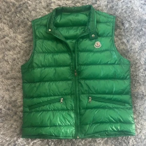 Grön dunväst från Moncler väst gui - Snygg grön dunväst från Moncler med klassisk logotyp på bröstet. Västen har dragkedja framtill och två snedställda fickor med dragkedja. Perfekt för lager-på-lager och ger en sportig look.