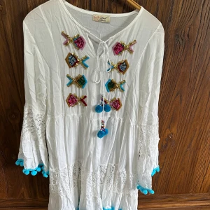 Vit bohemisk klänning med broderade detaljer - Vit klänning med bohemisk känsla, dekorerad med färgglada broderier och tofsar framtill. Har vida ärmar med spetsinslag och blå bollar längs ärmslut och fåll. Perfekt för sommarens varma dagar.