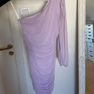 Ljuslila oneshoulder klänning - Säljer en snygg OANVÄND ljuslila oneshoulder klänning med lång ärm och draperad effekt. Klänningen har en tight passform.