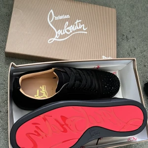 Svarta sneakers från Christian Louboutin med nitar - Säljer ett par svarta sneakers från Christian Louboutin med mattsvarta nitar på tån och klassisk röd sula. Skorna har snörning och är tillverkade i mocka och skinn. Kommer med originalkartong och dustbag. Har även kvitto