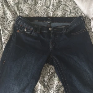 True Religion Joey Low Rise Flare Jeans - Mörkblå jeans från True Religion, modell Joey Low Rise Flare. Byxorna har låg midja, utsvängda ben och snygga detaljerade bakfickor med knappar och kontrastsömmar. Klassisk femficksmodell med logopatch bak. 