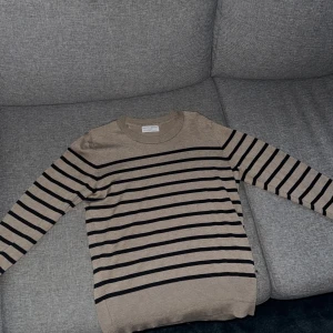 Beige och svart randig tröja från Holes & White - Säljer en beige långärmad tröja från Holes & White med svarta ränder. Tröjan har rund halsringning och dekorativa knappar vid ärmslutet. Super fin tröja men tyvärr inte ansvänt så mycket, endast använd några få gånger. Vet att den har någon jätte liten fläck någonstans men kunde inte hitta den, väldigt liten som sagt🩷 