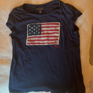 Mörkblå t-shirt med amerikansk flagga från Polo Ralph Lauren - Mörkblå t-shirt från Polo Ralph Lauren med en stor broderad amerikansk flagga på bröstet och röda RL-detaljer. På ärmen står det POLO i rött. Klassisk passform och mjuk bomullskvalitet.