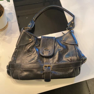 Svart axelväska i skinn med spänne - Säljer en svart axelväska i skinn med snygga detaljer som spänne och dragkedja. Väskan har både fram- och bakficka samt ett rejält handtag. Perfekt för dig som vill ha en stilren och praktisk accessoar.