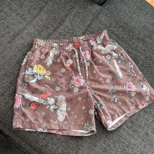 Bruna blommiga shorts  - Säljer ett par bruna shorts med färgglatt blommönster och stjärnor. Shortsen har snörning i midjan och är perfekta för sommaren. Mjukt och lätt material som känns skönt mot huden.