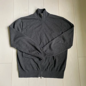Grå stickad kofta från Massimo Dutti - Säljer en stilren grå stickad kofta från Massimo Dutti med dragkedja framtill och hög krage. Perfekt för lager-på-lager och enkel att matcha med olika outfits. Mjuk och bekväm känsla tack vare det stickade materialet.
