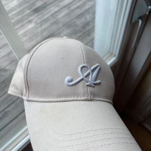 Beige keps från Axel Arigato - Snygg beige keps från Axel Arigato med vit broderad A-logga framtill. Klassisk böjd skärm och justerbar passform. Perfekt accessoar för en stilren och avslappnad look.