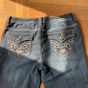 Mörkblå jeans med broderade bakfickor från Suko - Ett par mörkblå/ grå jeans från Suko med snygga broderade detaljer och nitar på bakfickorna. Köpta på vinted men passade tyvärr inte. Aldrig använda av mig. Färgen är lite mer blå irl.  Mått: innerben: 78cm ytterben: 102cm midja bred(mätt platt): 37cm midja höjd: 20cm