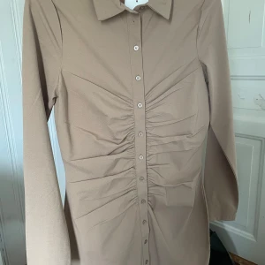 Beige skjortklänning med knappar från Vila - Säljer en beige skjortklänning från Vila i storlek XS. Klänningen har långa ärmar, krage och knappar hela vägen framtill. Snygg rynkdetalj längs knappraden som ger en cool form. Perfekt för dig som gillar stilrena och trendiga plagg.